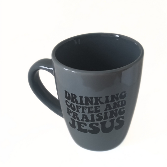 A gray ceramic mug with the text 'DRINKING COFFEE AND Praising Jesus' printed on the side.