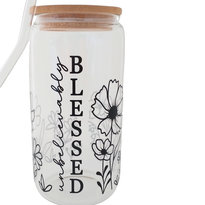 A clear glass reusable cup with black text and floral designs, a bamboo lid, and a glass straw.